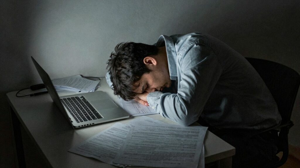 Exhausted person slumped over a desk in a dark room.