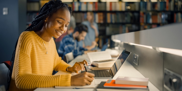 University Library: Gifted Black Girl uses Laptop, Writes Notes for the Paper, Essay, Study for Class Assignment. Diverse Multi-Ethnic Group of Students Learning, Studying for Exams, Talk in College
