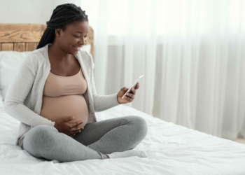 Smiling Black Pregnant Woman Sitting With Smartphone On Bed At Home, Looking At Cellphone Screen With Excitement, Happy African Lady Browsing Pregnancy App Or Received Mobile Offer, Copy Space