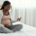 Smiling Black Pregnant Woman Sitting With Smartphone On Bed At Home, Looking At Cellphone Screen With Excitement, Happy African Lady Browsing Pregnancy App Or Received Mobile Offer, Copy Space
