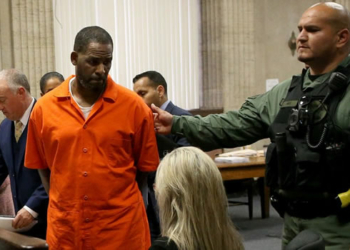 R Kelly in a court room escorted by a guard